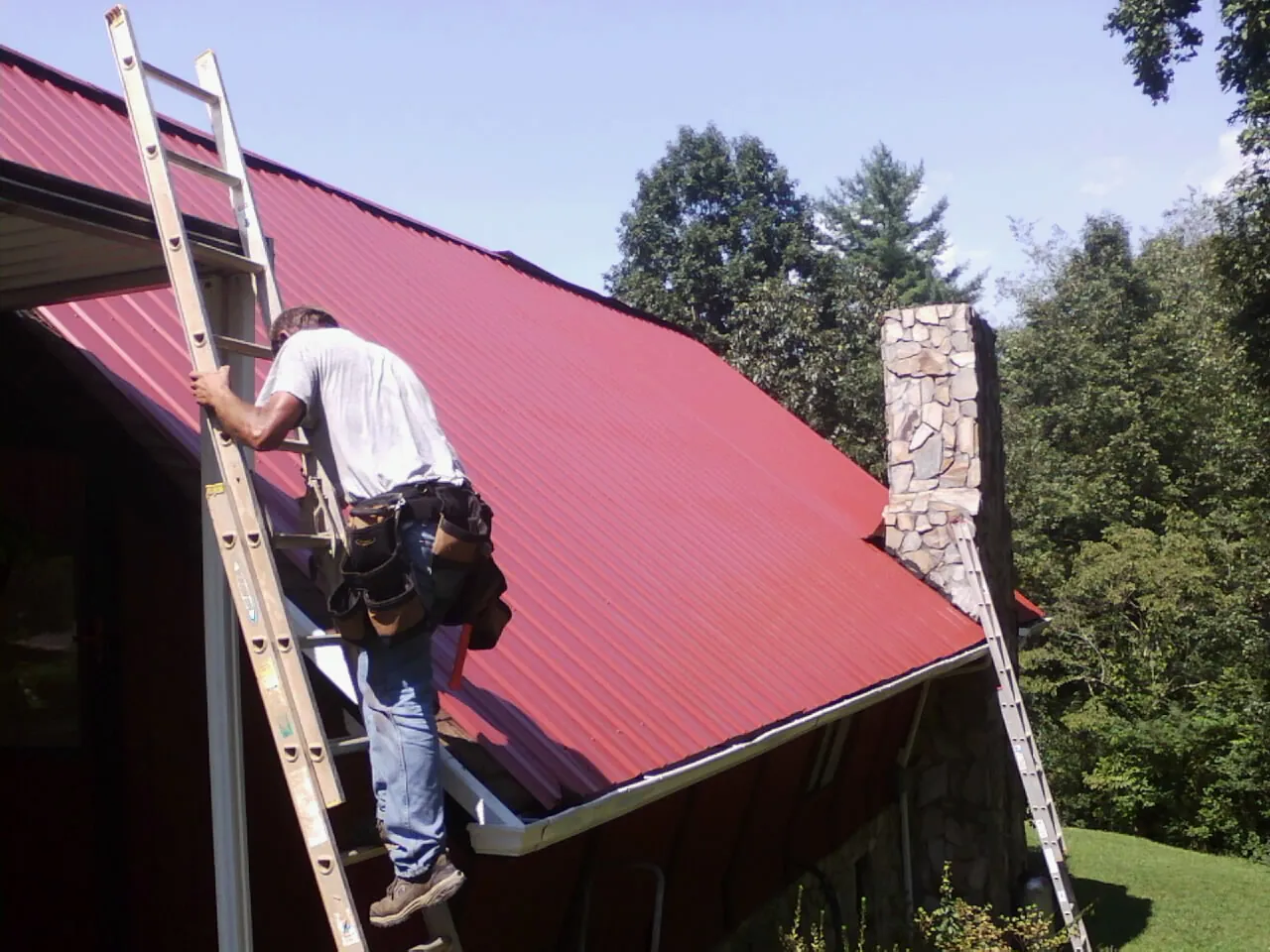 Roofer on a ladder beside a red metal roof and stone chimney — Soffit & Fascia Repair by Roofing Brothers in Great River